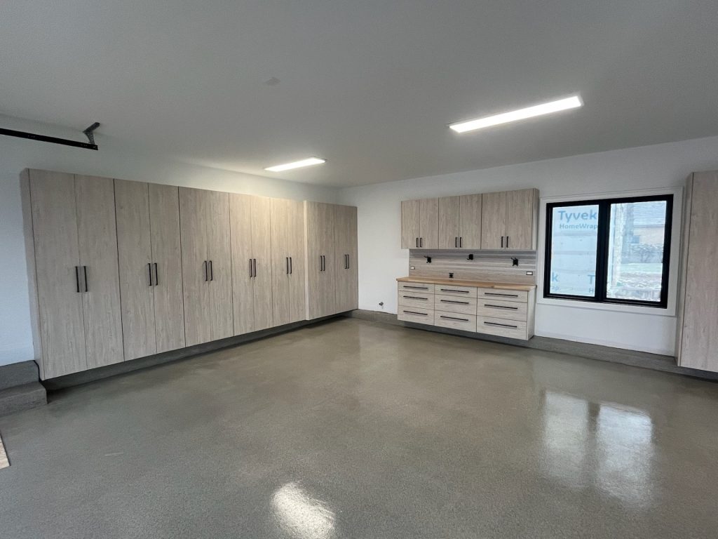 A spacious, clean garage with light wooden cabinets on one wall and a workbench with drawers under a window on another wall. The floor is polished concrete, and the room is well-lit with ceiling lights.