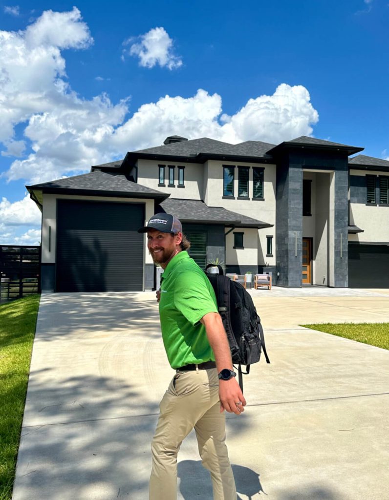 A man in a green shirt, beige pants, and a cap walks on a driveway in front of a modern two-story house under a blue sky with clouds. He carries a black backpack.