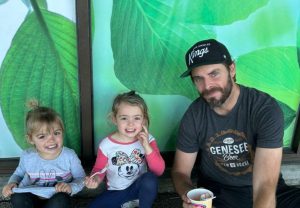 A man sitting with two young children in front of a large green leaf mural. The children are smiling and holding spoons, while the man holds a cup.