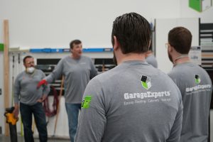 Group of people wearing "GarageExperts" shirts, listening to a speaker in a workshop setting with tools and materials in the background.