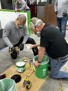 Two people wearing masks mix white paint in green buckets using drills, in a workshop setting.