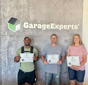 Three people stand in front of a wall with the GarageExperts logo, each holding a certificate.