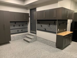 Garage interior with gray cabinets, wall-mounted storage panels, and a workbench. Floor is speckled gray, and there is a small set of steps leading to a door.